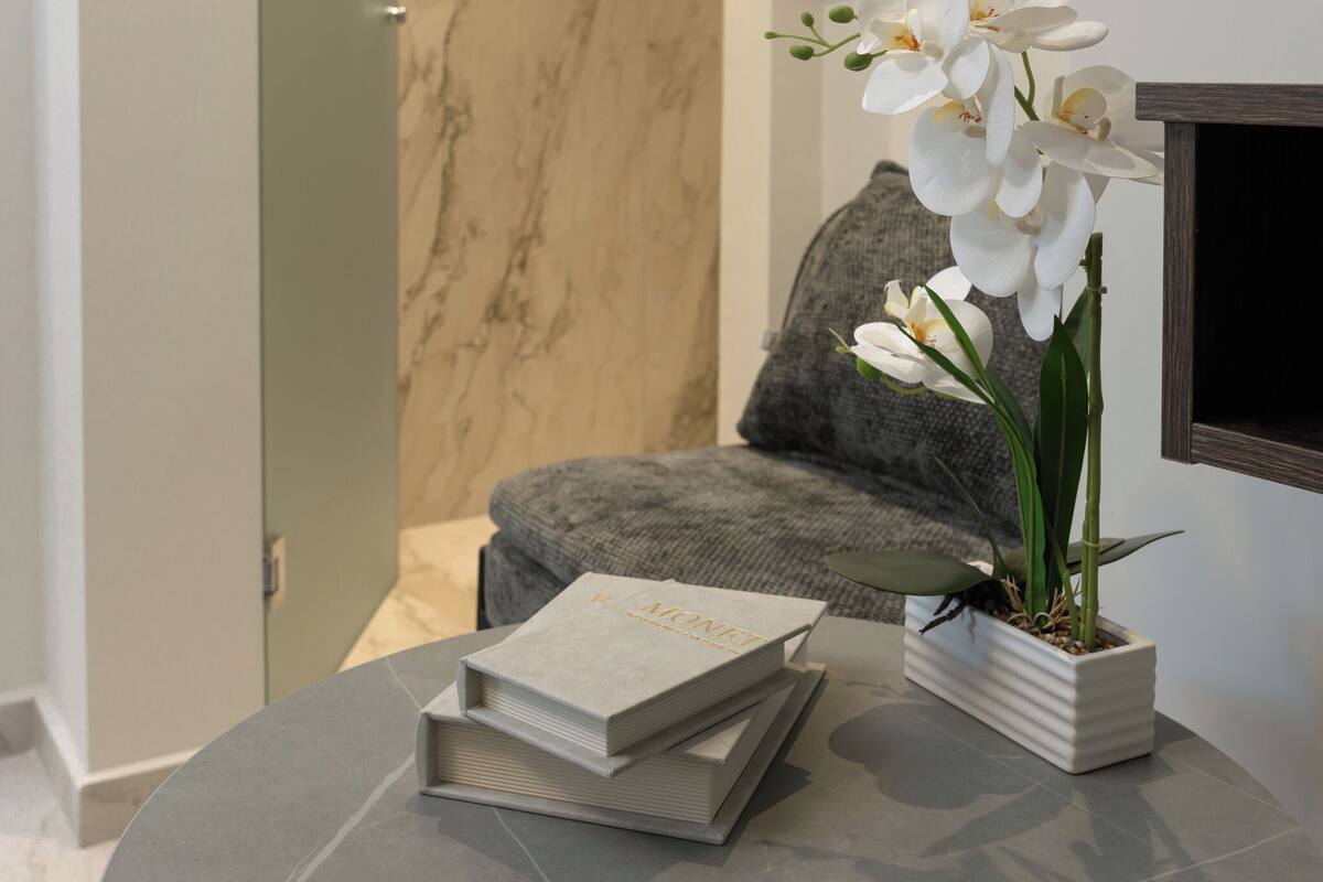 Una habitación con suelo de mármol, un sofá gris, una orquídea blanca en maceta blanca y una pila de libros sobre una mesa redonda.