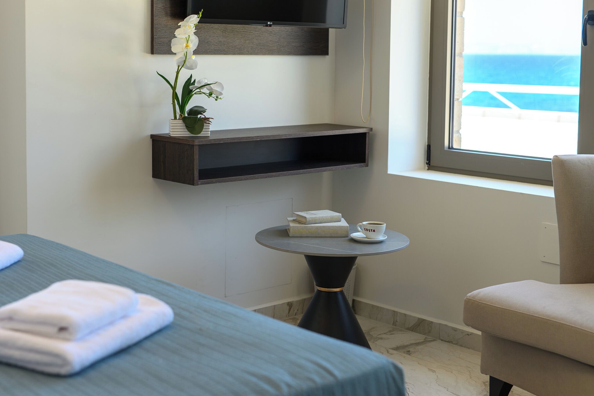 Une chambre d’hôtel avec un lit, une petite table ronde sur laquelle se trouvent une tasse et un livre, une étagère avec une plante et une fenêtre donnant sur la mer.