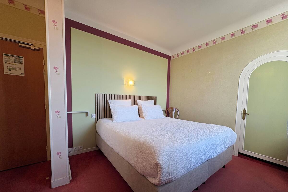 Una cama bien hecha en una habitación con una puerta a la izquierda y una lámpara de pared sobre la cama.