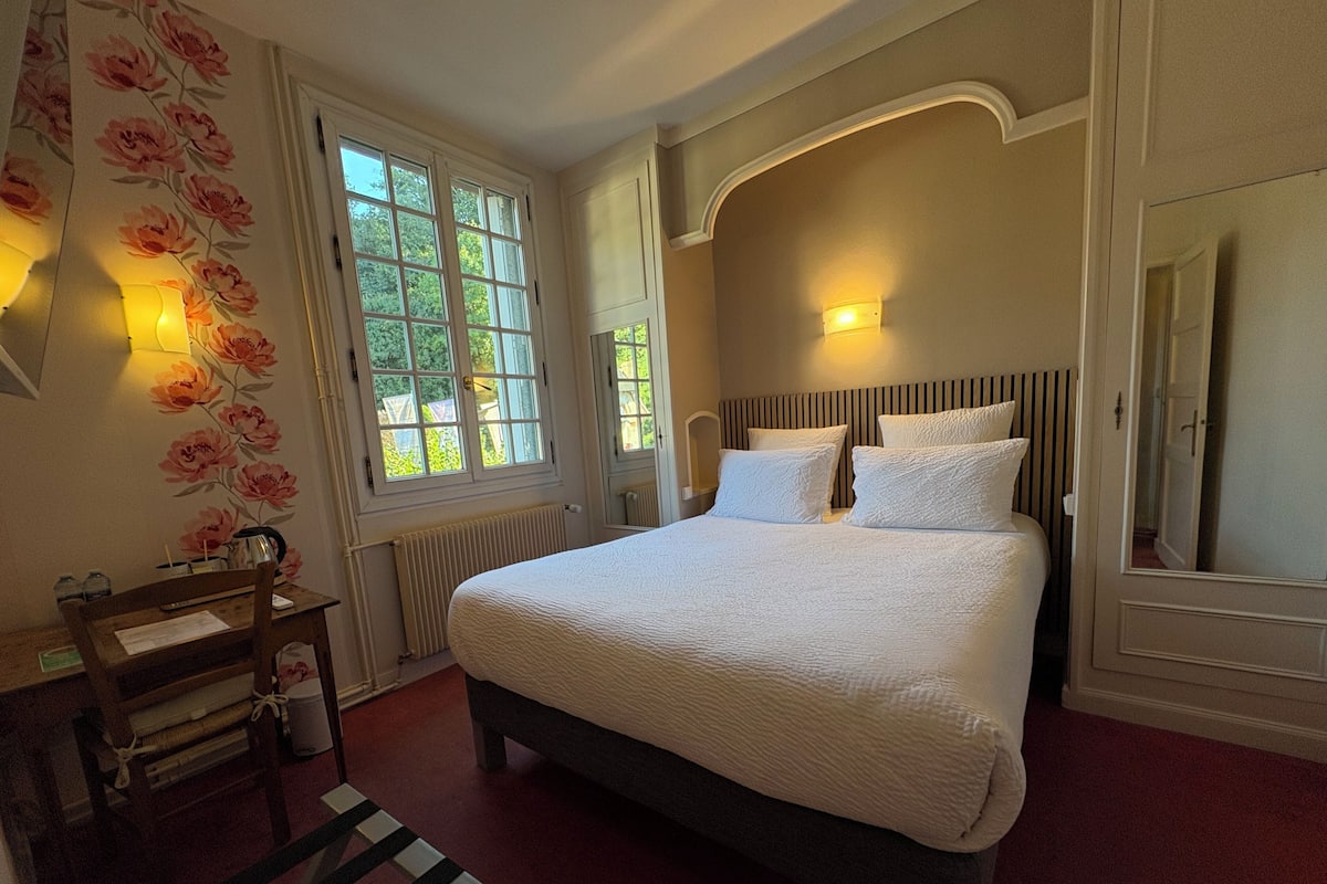 Un dormitorio con una cama grande, una mesita de noche de madera, un radiador y una ventana con papel tapiz floral.