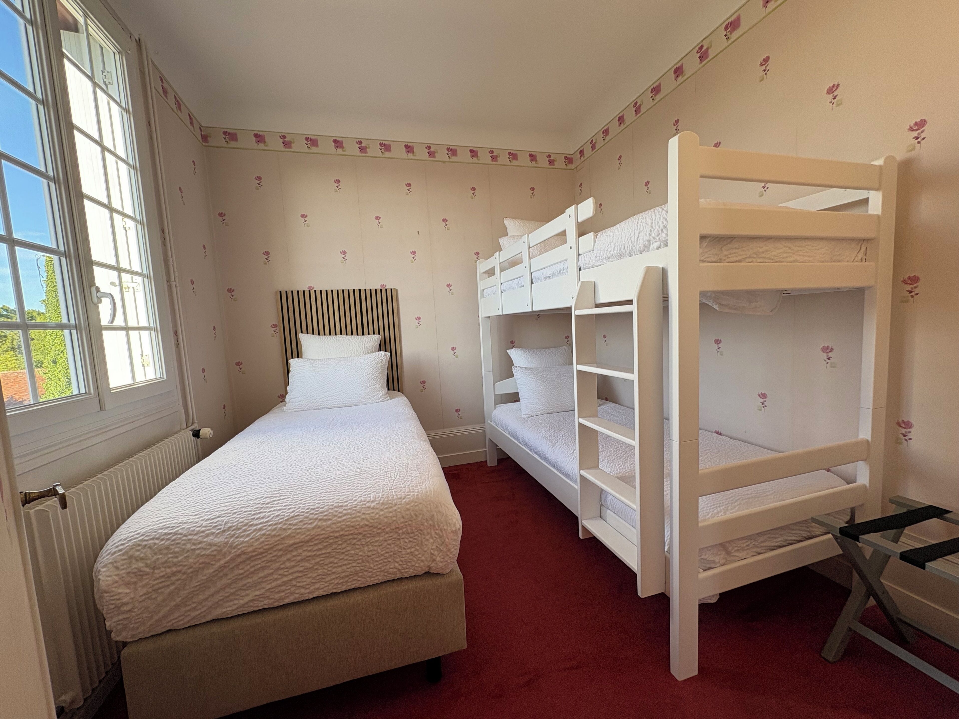 Une chambre avec un lit simple, deux lits superposés, un radiateur et un papier peint à motifs floraux.