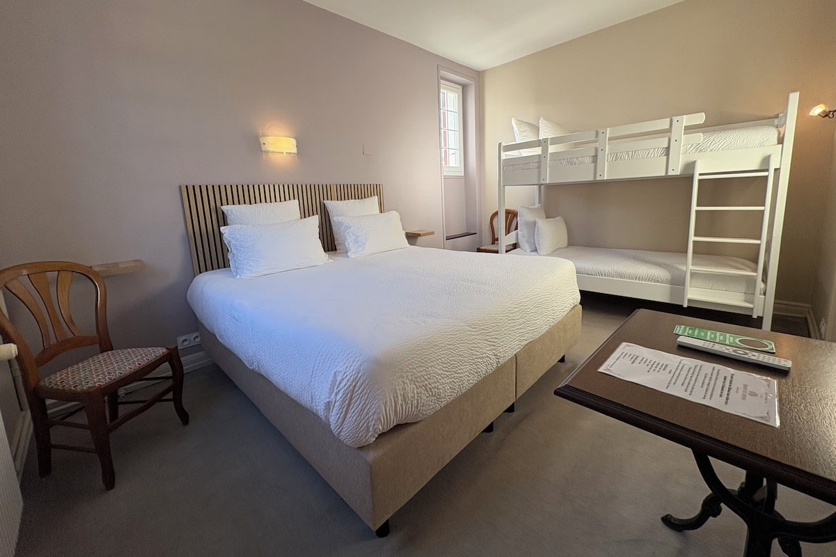 Habitación de hotel con una cama individual, una silla de madera, una mesita con un control remoto y una cama litera.