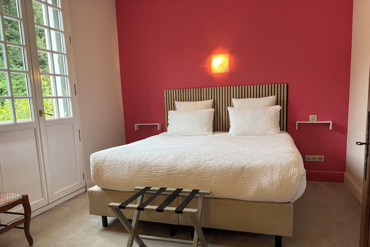Un dormitorio con una cama, una mesita de noche, una silla, un ventanal y una lámpara de pared.