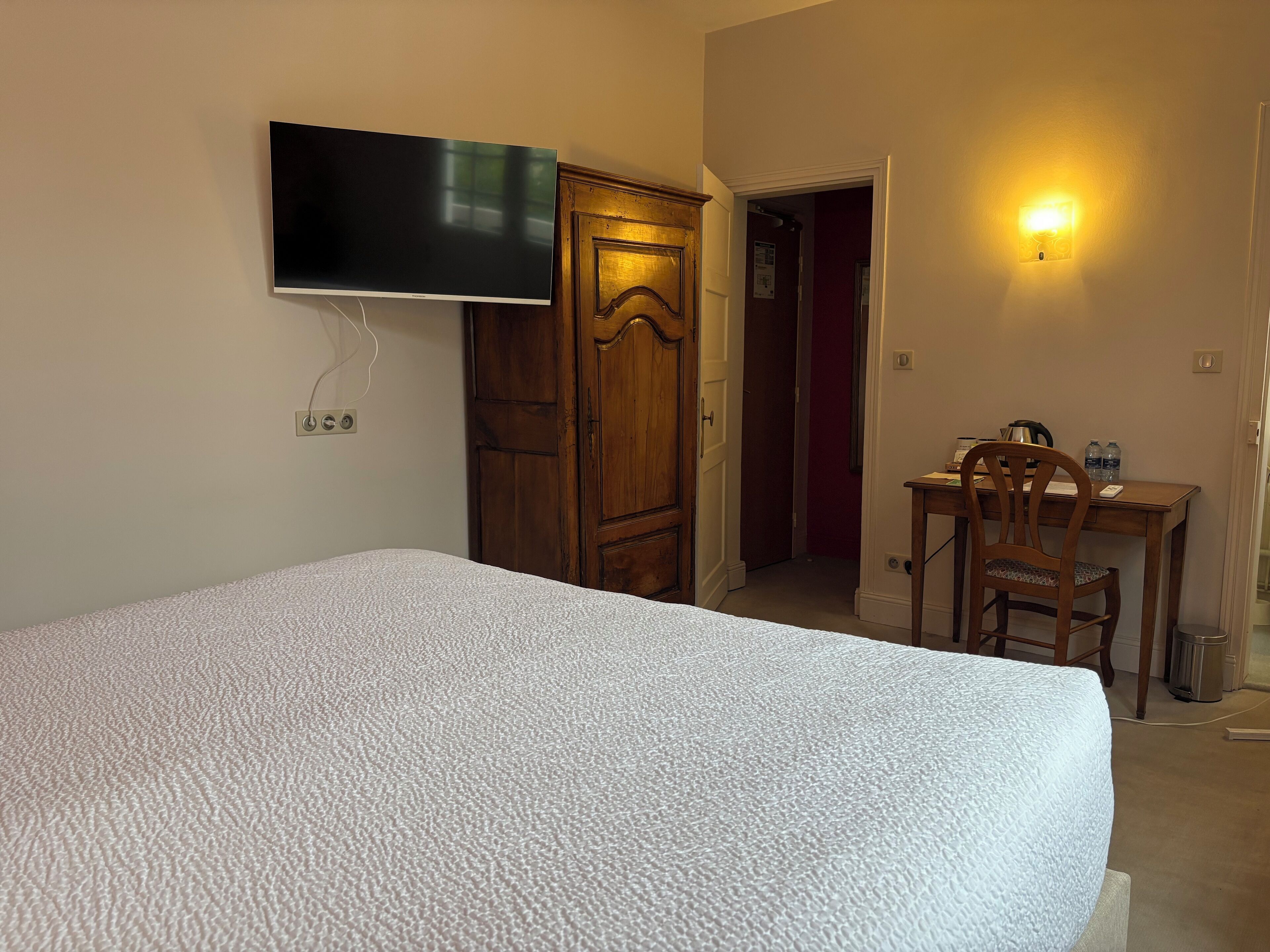 A bedroom with a bed, a TV mounted on the wall, a wooden wardrobe, and a desk with a chair.