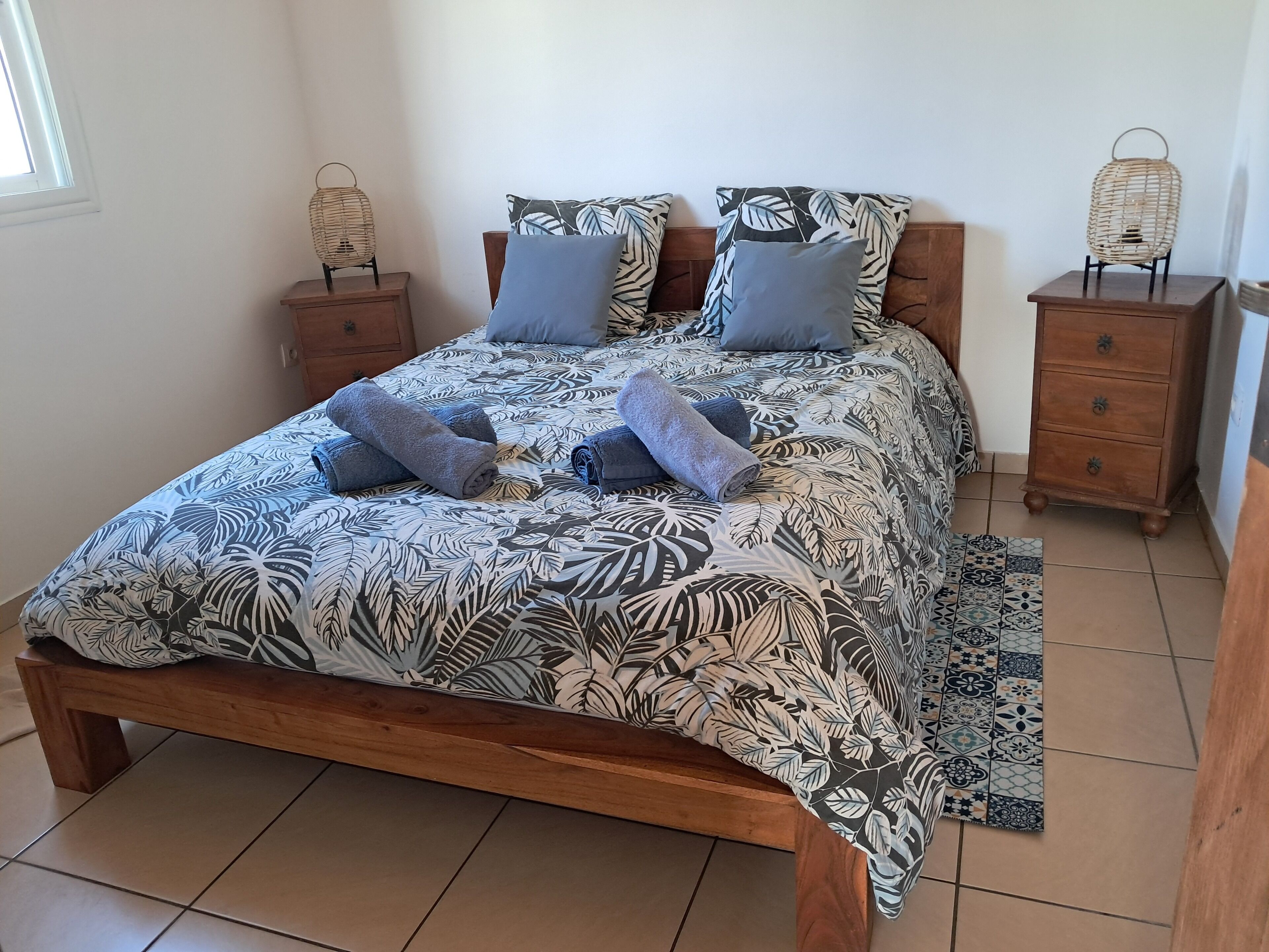 Ein ordentlich bezogenes Bett mit einem blattgemusterten Überwurf, zwei Nachttische mit Korbtischlampen und ein gemustertes Teppich.