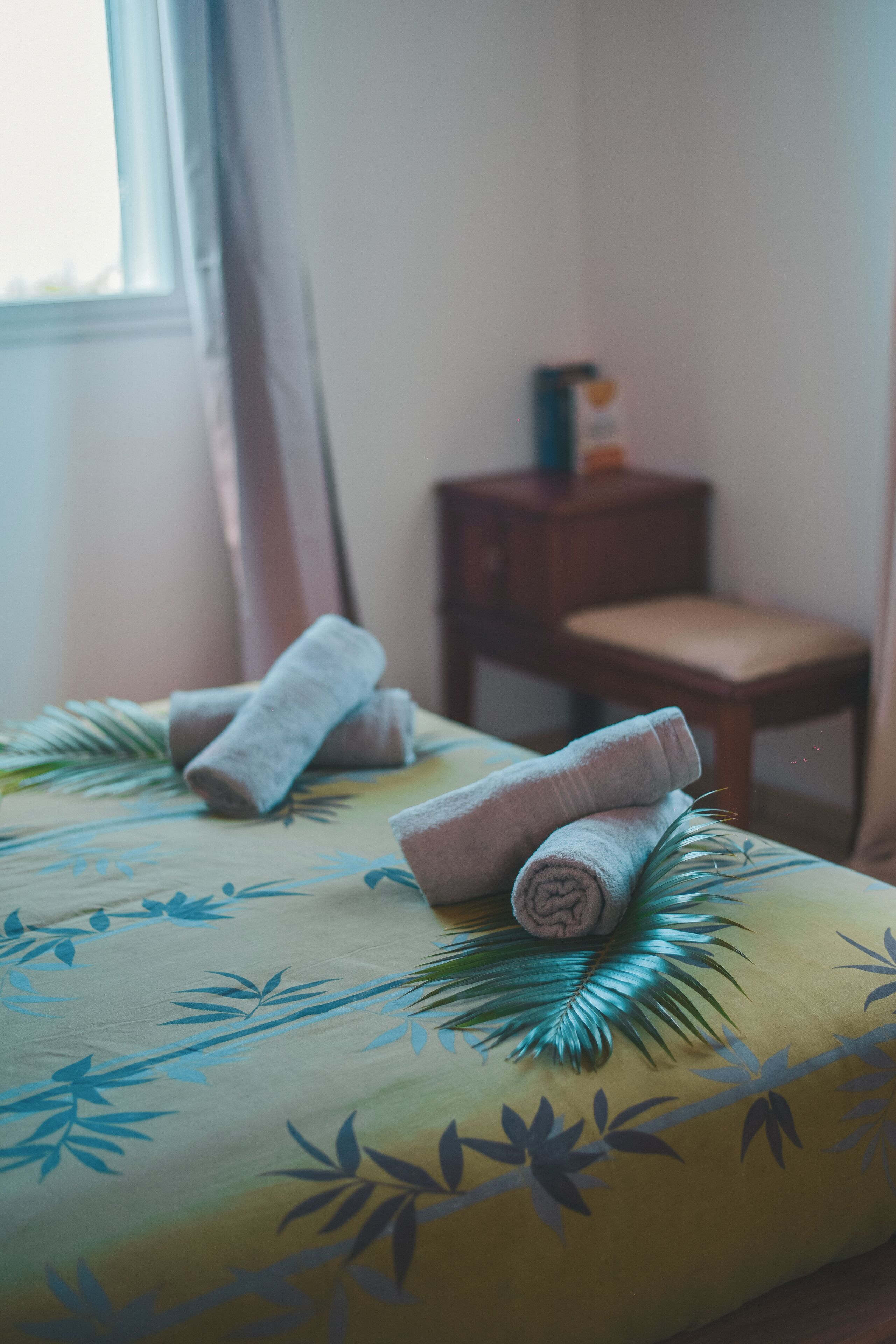 A bed with rolled towels and a palm leaf design, a wooden nightstand, and a chair in a room with a window.