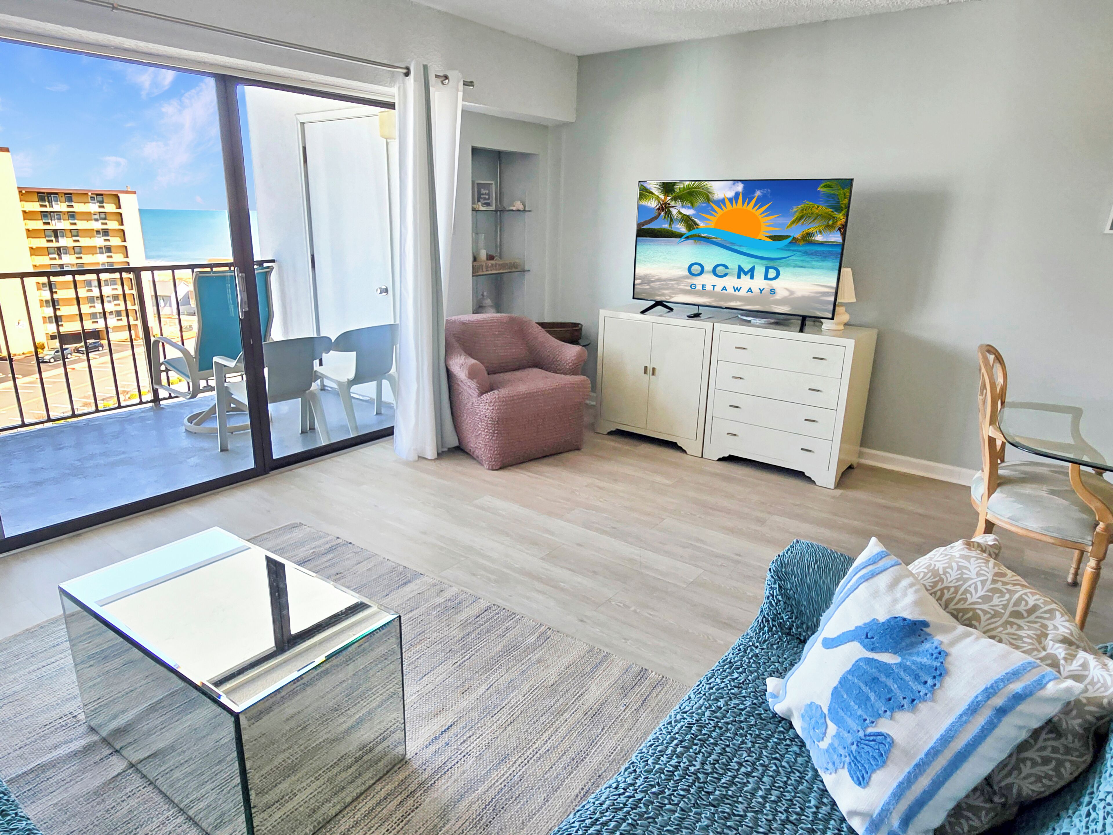 Ein Wohnzimmer mit Glastisch, einer Couch mit dekorativen Kissen, einem Fernseher auf einer Anrichte und einem Balkon mit Blick auf das Meer und Gebäude.