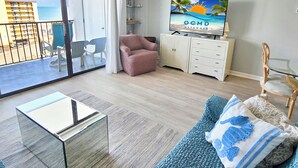 Ein Wohnzimmer mit Glastisch, einer Couch mit dekorativen Kissen, einem Fernseher auf einer Anrichte und einem Balkon mit Blick auf das Meer und Gebäude.