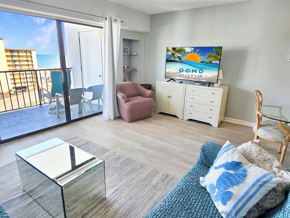A living room with a glass coffee table, a sofa with decorative pillows, a television on a cabinet, and a balcony with a view of the ocean and buildings.