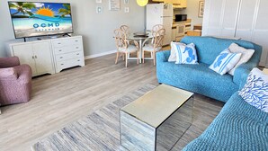 Ein Wohnzimmer mit Fernseher, Sofa, Sesseln und einer Küche im Hintergrund.