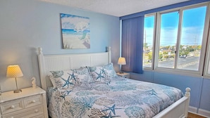 A bedroom with a bed, nightstands, a window with a view, and a painting on the wall. - Oceanfront 3rd Floor Condo with Pool just steps from the Atlantic Ocean! Quay #301 (Ocean City)