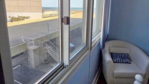 A room with a large window overlooking a parking area and a building. There is a small seating area with a cushion and a welcome sign. - Oceanfront 3rd Floor Condo with Pool just steps from the Atlantic Ocean! Quay #301 (Ocean City)
