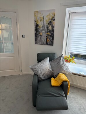 Une pièce avec un canapé gris, un tableau représentant la Tour Eiffel, une fenêtre avec des stores et un radiateur.