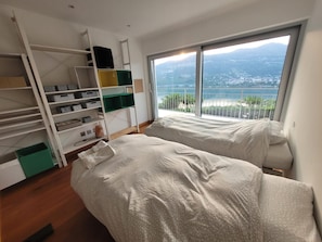 Een slaapkamer met twee bedden, een inbouwkleerkast en een groot raam met uitzicht op een kustlandschap.