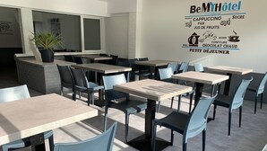 A hotel dining area with multiple tables and chairs, a potted plant, and a wall with a menu listing various food and drink options. - BE MYHOTEL (Évreux)