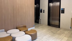 Ένας σύγχρονος χώρος ανελκυστήρων με επένδυση τοίχου από ξύλινα πάνελ, μια περιοχή με καθίσματα και στρογγυλά σκαμπό και ένα δάπεδο με πλακάκια.