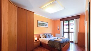 A bedroom with a wooden wardrobe, a bed with a blue blanket, a nightstand, and a window with curtains.