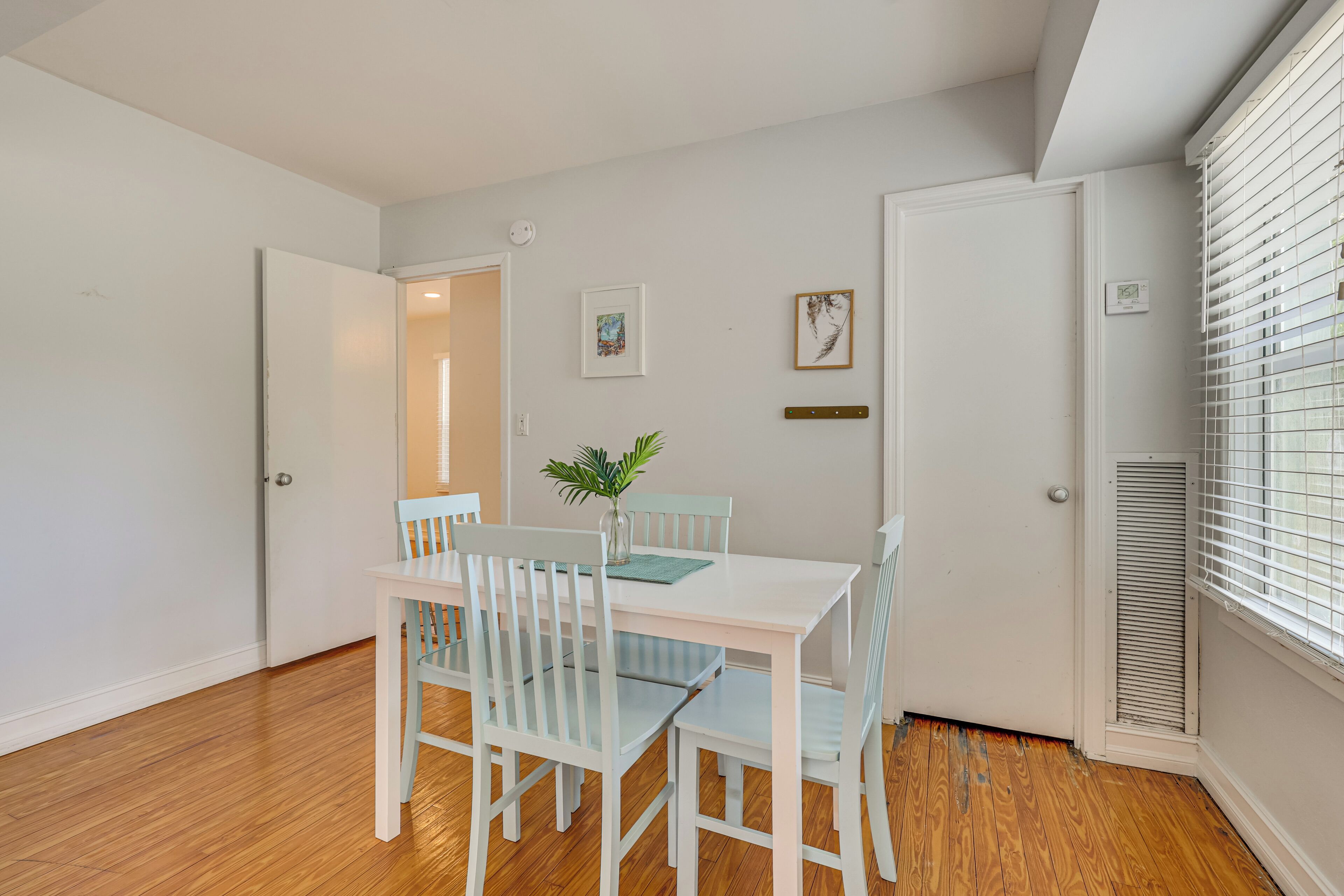 A dining area with a white table and chairs, a vase with a plant, and a window with blinds.
