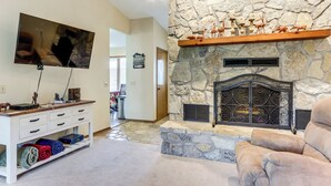 A living room with a stone fireplace, a mounted television, and a plush armchair. - Holiday Island Family Home on 13th Green! (Eureka Springs)