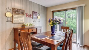 A dining area with a wooden table, chairs, and a vase of flowers. There are wall decorations and a window with curtains. - Holiday Island Family Home on 13th Green! (Eureka Springs)
