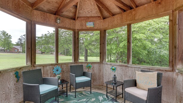 A covered porch with wicker chairs and a table, large windows, and a view of a green landscape. - Holiday Island Family Home on 13th Green! (Eureka Springs)