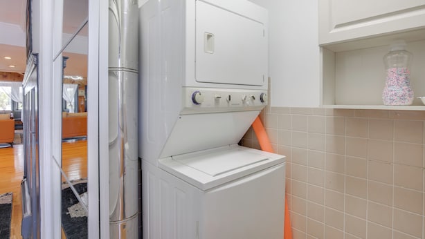 Une petite buanderie moderne et fonctionnelle, dotée d’un lave-linge et d’un sèche-linge intégrés, d’une petite étagère où sont exposés des objets décoratifs, et qui offre une vue sur l’espace de vie.