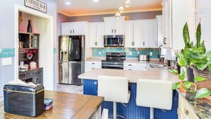 Una cocina con armarios blancos, una isla central con taburetes de bar, electrodomésticos de acero inoxidable y un letrero que dice 'Coffee Bar'.