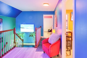 A brightly colored hallway with a red chair, a staircase with a wooden railing, and a door leading to a room with a view of the outdoors. - Island Vibes: Home w/ Fire Pit in Murfreesboro! (Murfreesboro)