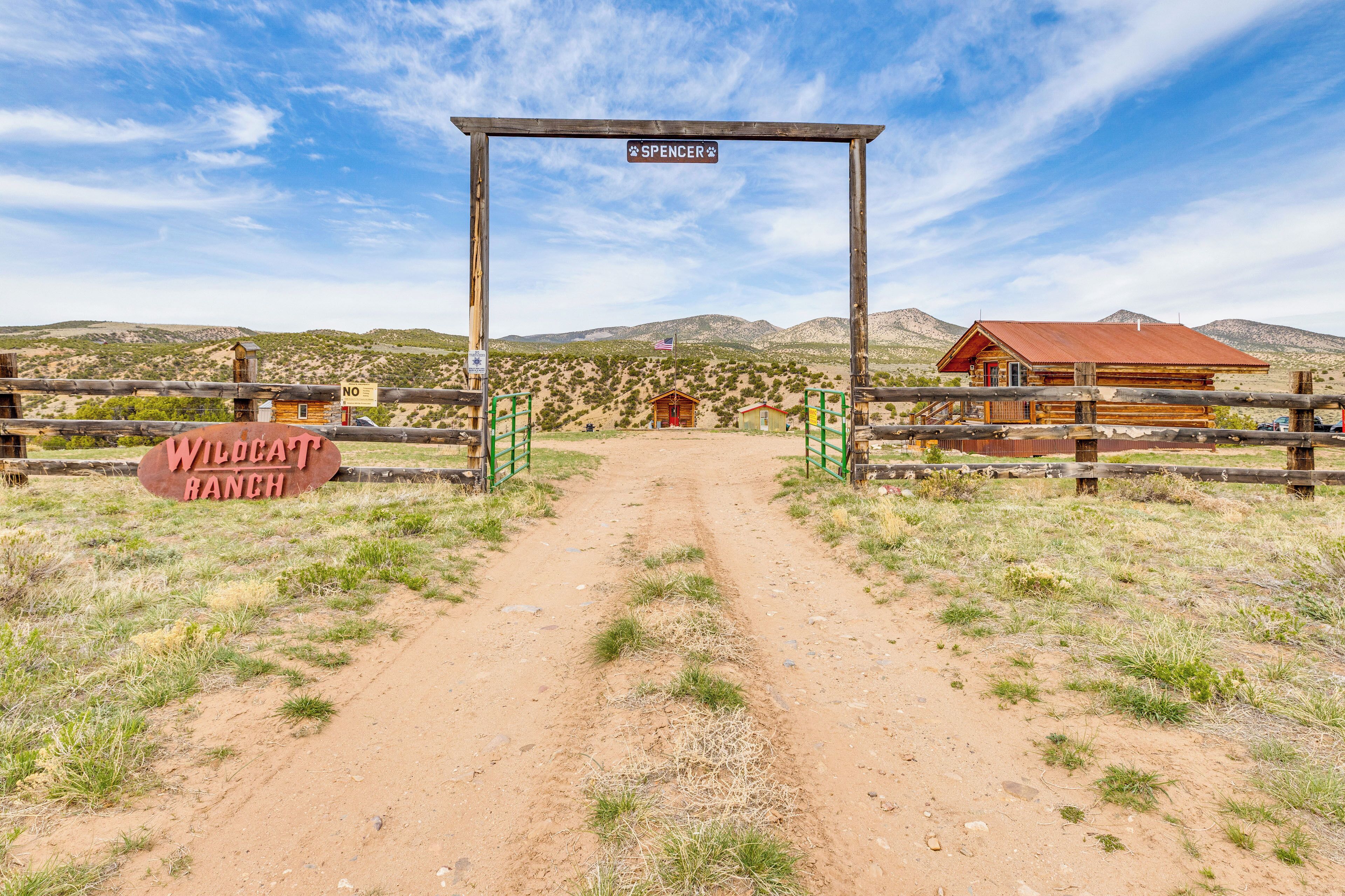 Un arco de madera con un letrero que dice "Spencer" y "Wildcat Ranch" que conduce a un edificio rústico con techo rojo.