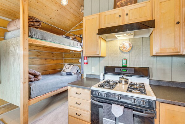 A compact kitchen with a stove, wooden cabinets, and a bunk bed with blankets. - Sunset & Mtn Views: Rustic Colorado Retreat! (Maybell)