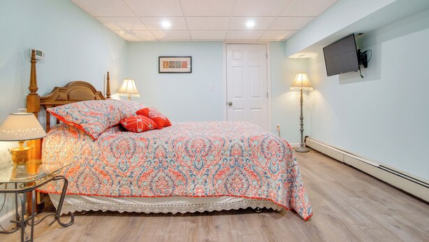 A bedroom with a bed, bedside lamps, a wooden headboard, a TV mounted on the wall, and a door. - Patio + Pond View: Serene Cape Cod Escape (Mashpee)