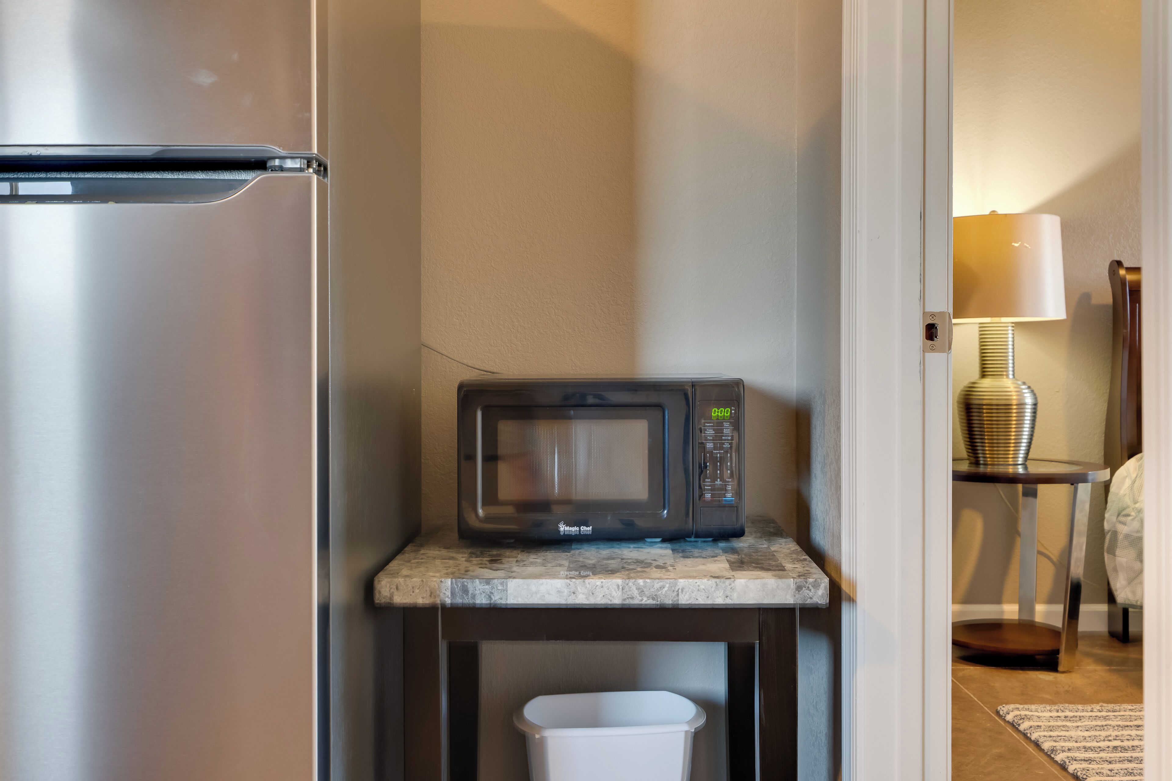 A compact kitchenette with a stainless steel refrigerator, a microwave on a small marble countertop, and a white trash bin below.