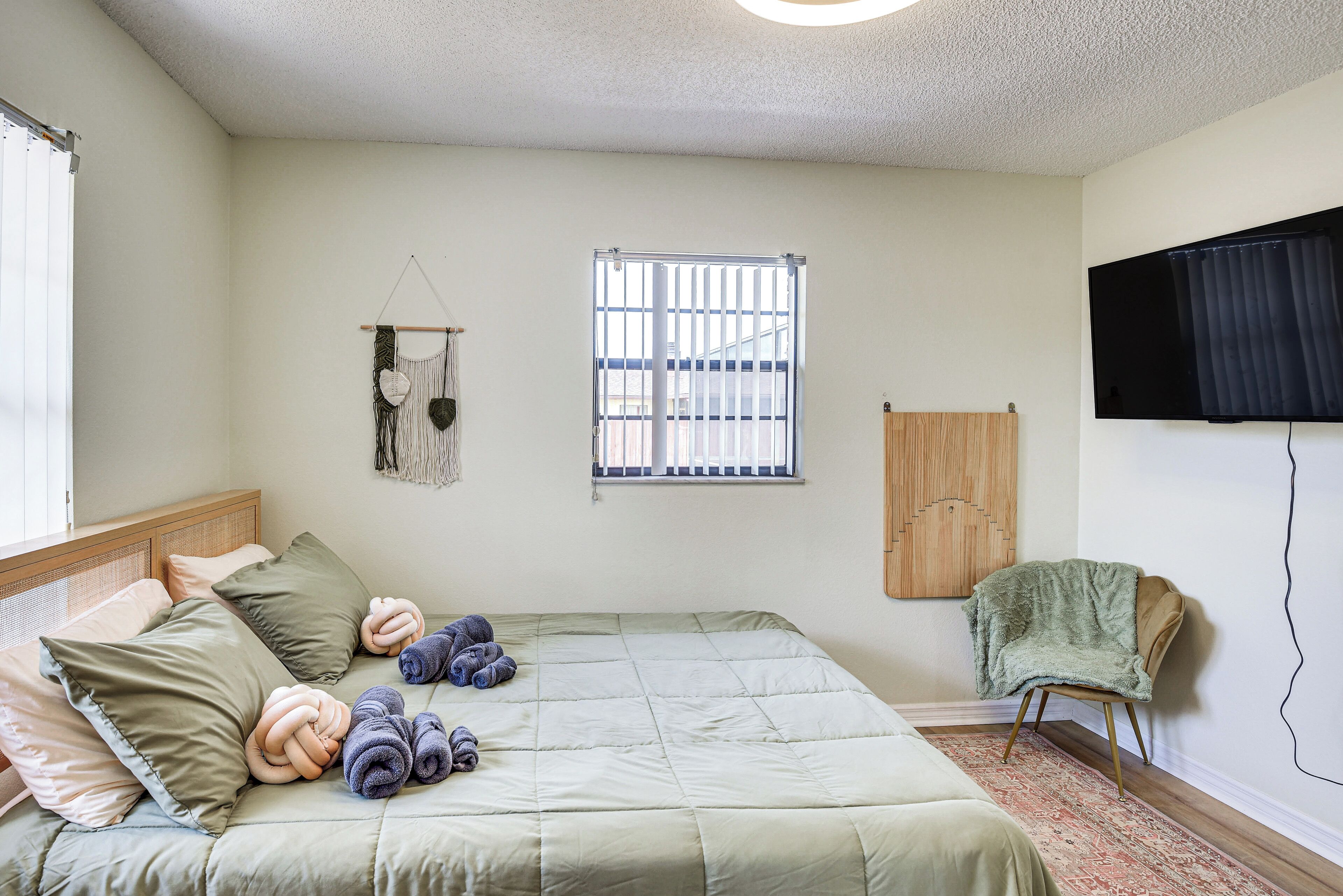 A neatly arranged bedroom with a bed, a chair, a window, and a wall-mounted TV.