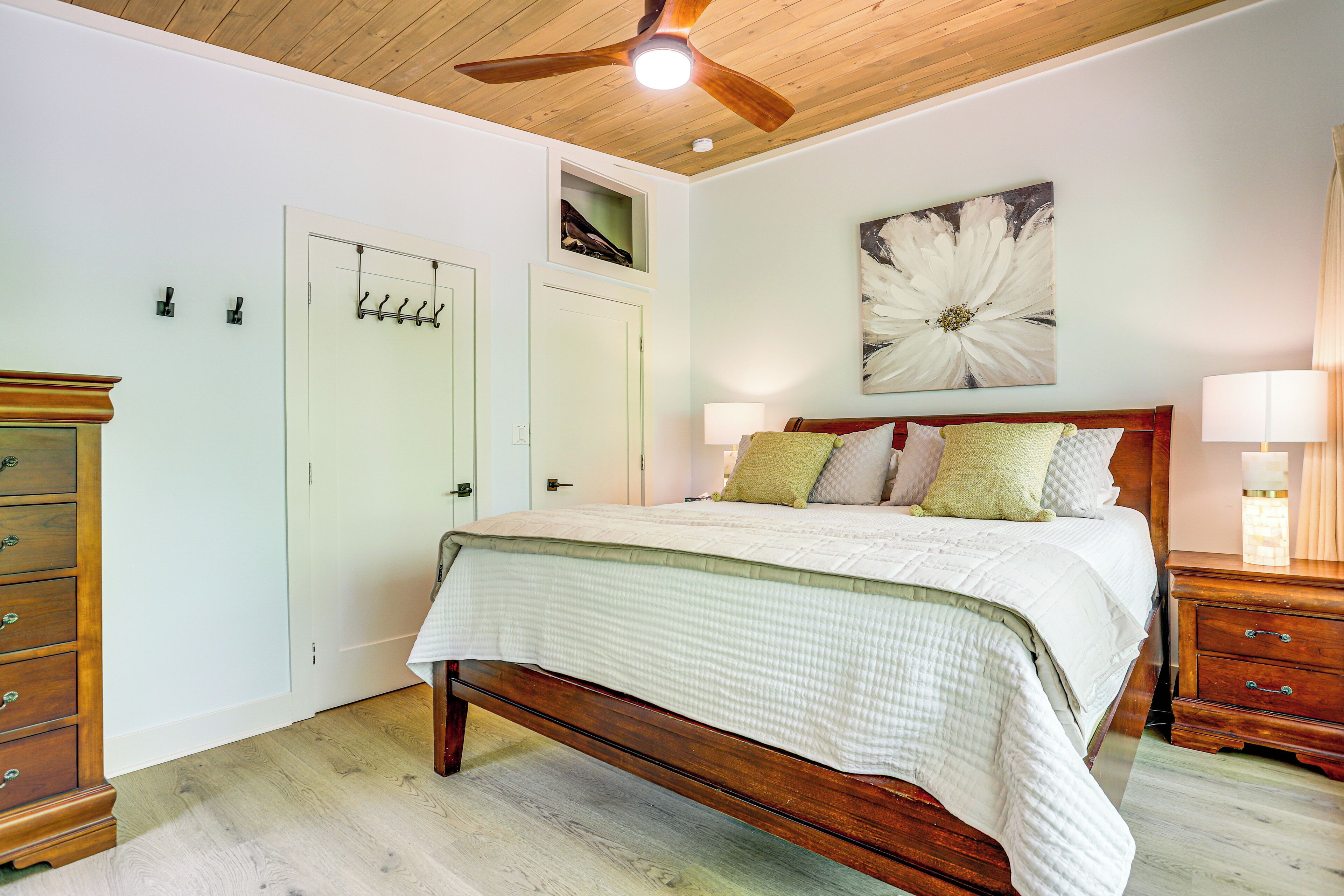 Une chambre à coucher avec un lit en bois, un ventilateur de plafond, un grand tableau représentant des fleurs et une table de chevet avec une lampe.
