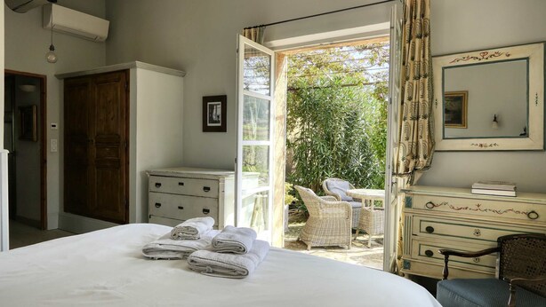 Une chambre à coucher comprenant un lit, une commode, une chaise et une porte ouverte donnant sur un balcon aménagé avec du mobilier d’extérieur.