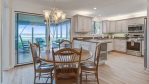 A modern kitchen with white cabinets, a dining area with wooden chairs, and a large window overlooking a patio. - Kiawah Exclusives | 165 Marsh Hawk Lane (Kiawah Island)