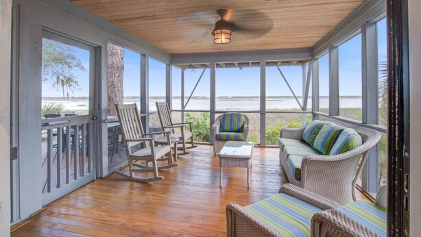 A covered outdoor area with wooden flooring, wicker furniture, and large windows offering a view of the beach. - Kiawah Exclusives | 165 Marsh Hawk Lane (Kiawah Island)