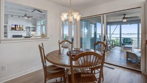 A dining area with a round wooden table and chairs, a chandelier, and a view through glass doors to an outdoor patio with seating. - Kiawah Exclusives | 165 Marsh Hawk Lane (Kiawah Island)