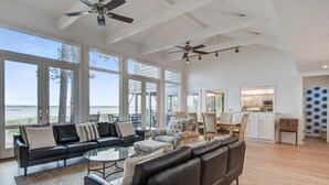 A spacious living area with a large black leather sofa, a glass coffee table, and a view of the ocean through large windows. There are ceiling fans, a kitchen area, and a dining table visible. - Kiawah Exclusives | 165 Marsh Hawk Lane (Kiawah Island)