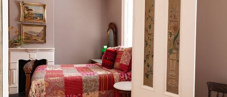 A bedroom with a large bed, a small table, and a chair. There are two paintings on the wall, a mirror, and a window with curtains.
