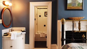 A bathroom with a white sink, a mirror, and a towel rack. There is a doorway leading to a shower area with a white tiled wall. A painting hangs on the wall above the doorway. - Jesse James - Historic Hotel (Cimarron)