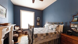 A bedroom with a large bed, a wooden dresser, a chair, a fireplace, and a ceiling fan. - Jesse James - Historic Hotel (Cimarron)