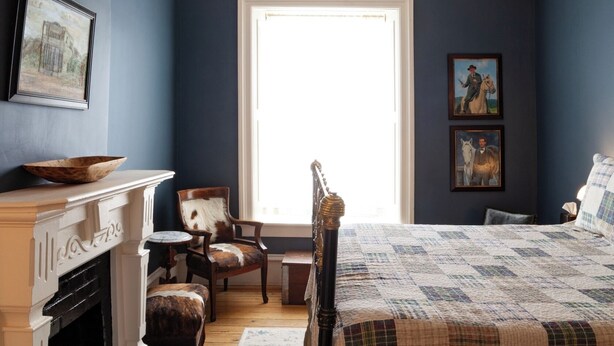 A bedroom with a large window, a fireplace, a bed with a quilt, a chair, and two framed pictures on the wall. - Jesse James - Historic Hotel (Cimarron)