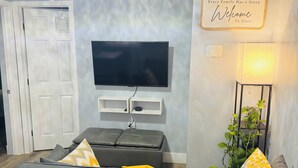 A living room with a grey sofa, a flat-screen TV mounted on the wall, a small table with a lamp, and a sign that says "Every Family Has a Story Welcome To Our Home."