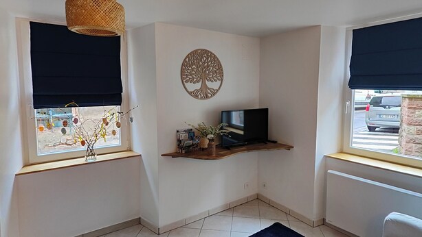 Ein kleiner, gut beleuchteter Raum mit einem großen Fenster, einem an der Wand befestigten Regal mit Dekorationen, einem dunkelblauen Teppich und einem weißen Sofa.