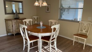 A dining room with a wooden table, white chairs, a chandelier, a mirror, and a window with blinds.