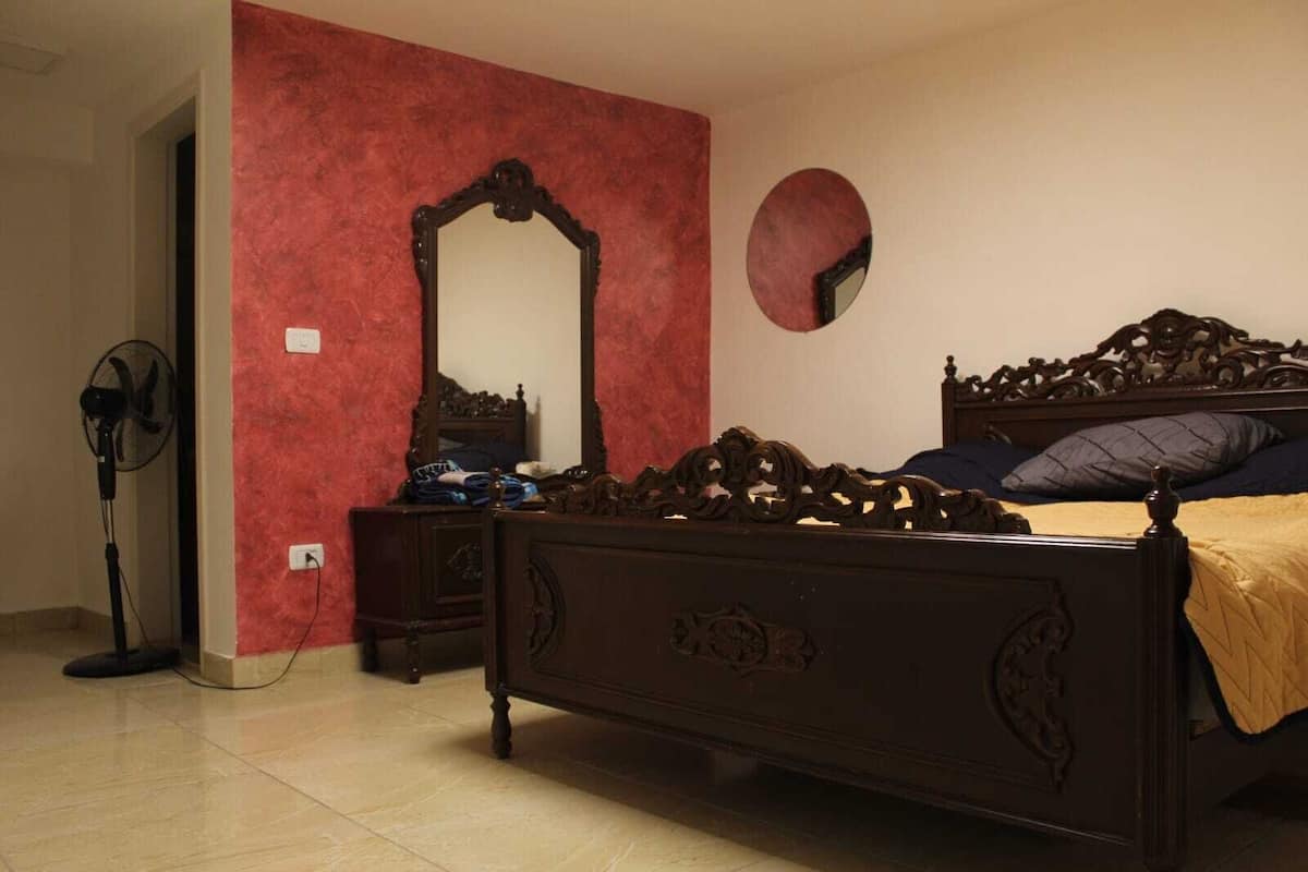 A bedroom with a wooden bed, a dresser, a mirror, and a standing fan.