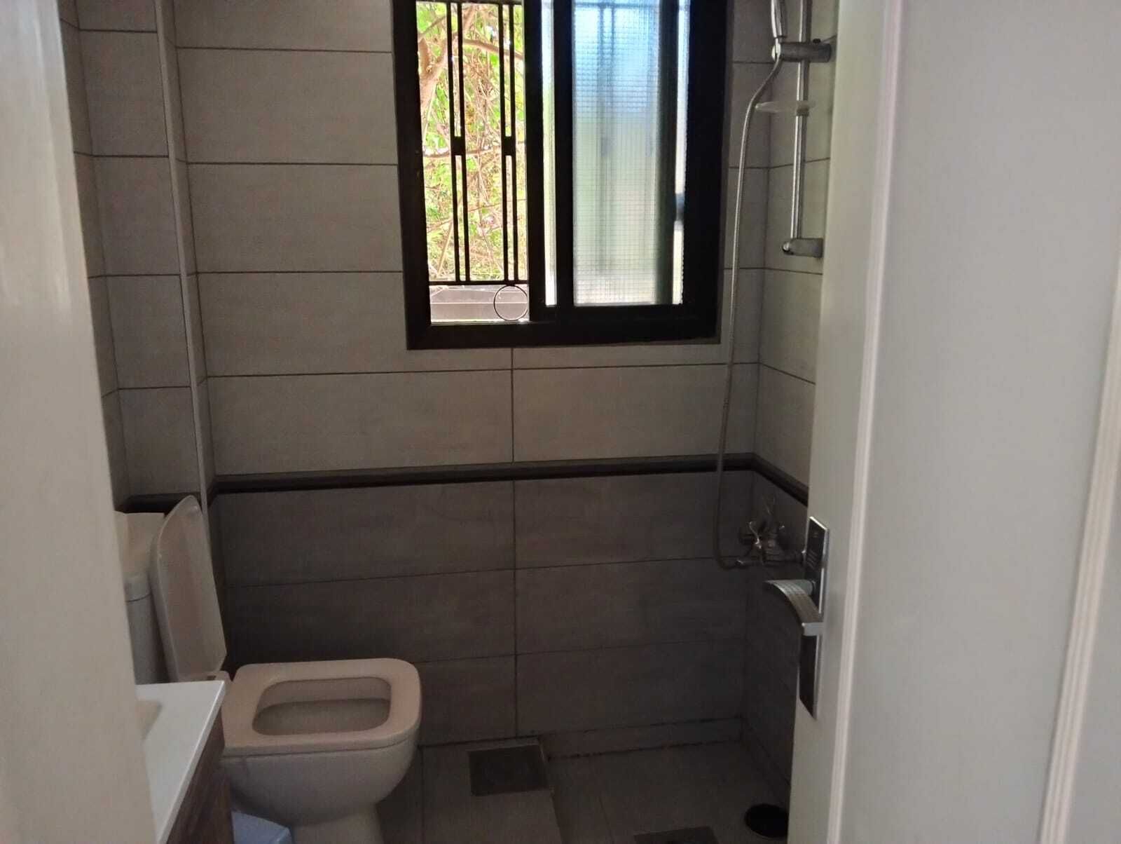 Een klein betegeld badkamertje met toilet, een raam met tralies en een douchehoek.