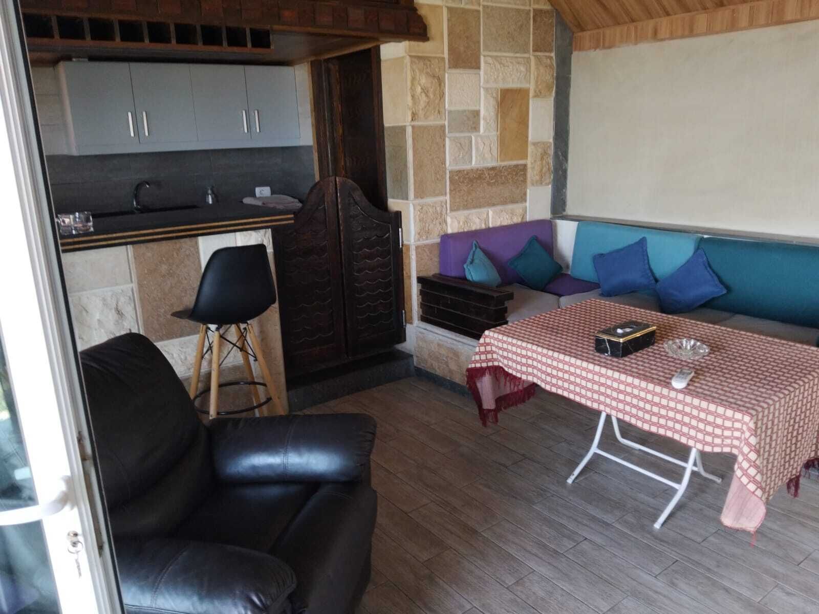 Un rincón al aire libre acogedor con una mesa cubierta por un mantel a cuadros, una silla de cuero negro y un muro de piedra de fondo.