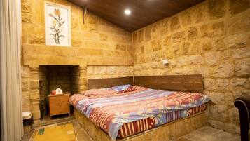 Quarto com parede de pedra, cabeceira de madeira, cama com colcha florida, mesa de cabeceira e lareira.